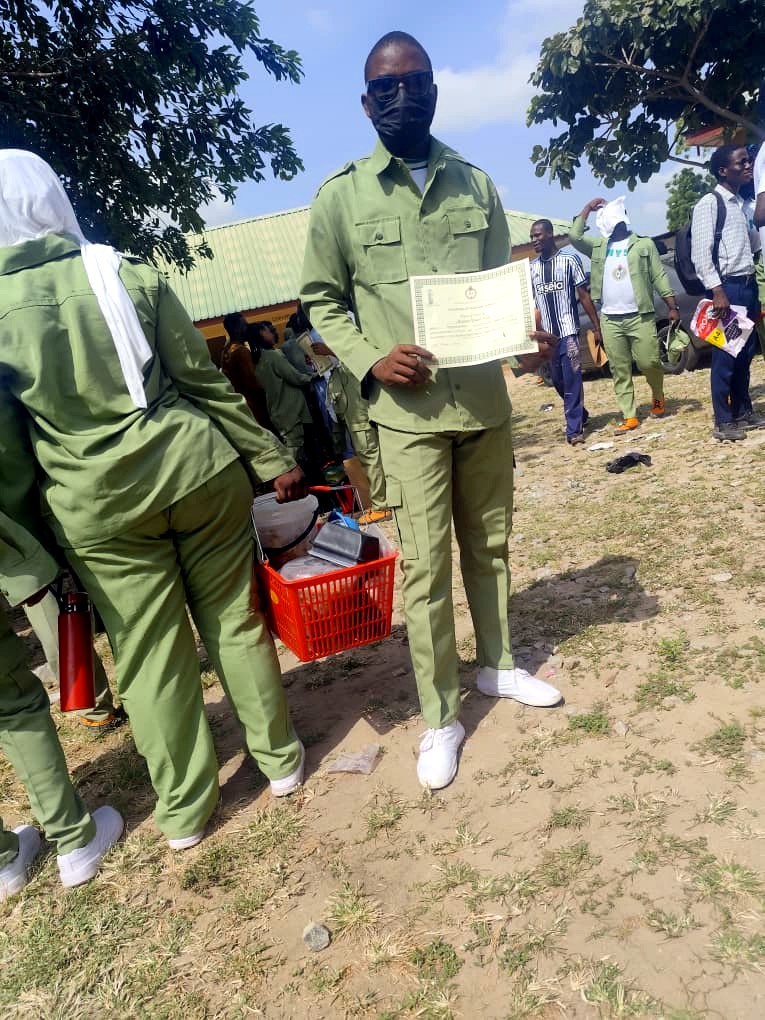Rasheed Taiwo standing in full NYSC uniform displaying his Certificate of National Service